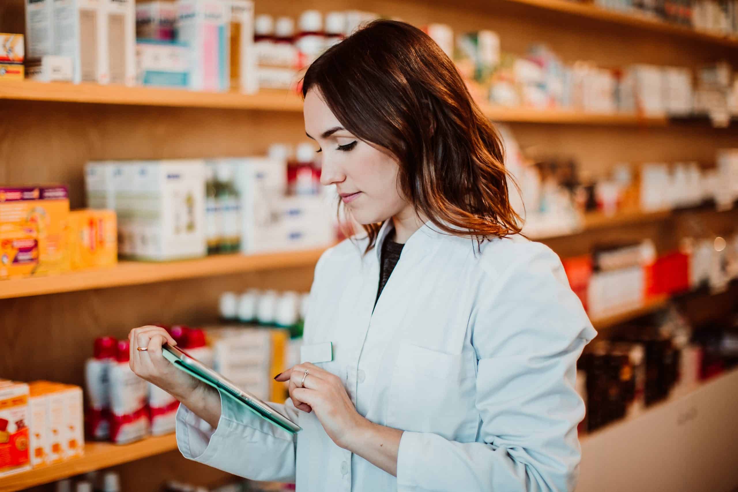 Eine Frau im weißen Kittel betrachtet ein Produkt vor einem Regal mit Arzneimitteln und Gesundheitsprodukten